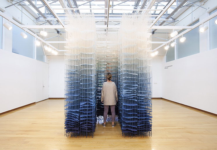 large hanging paper installation with white at top grading to blue at the bottom hanging in the center of a gallery; a person stands in the middle of it, facing away from the camera