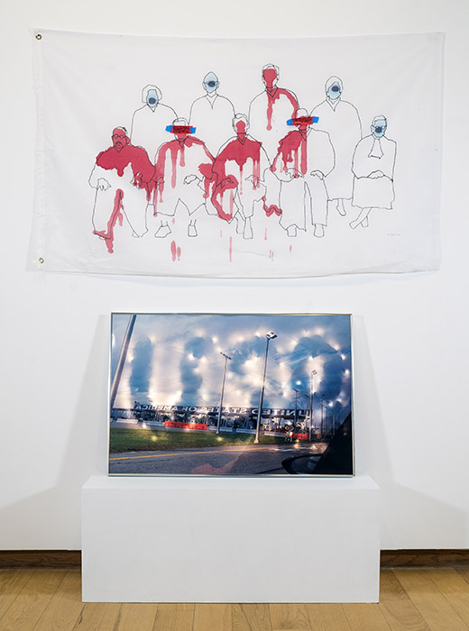 white flag with line drawing of Supreme Court justices, some splattered with red paint; on a low pedestal below, photo of the US/Canada border crossing with USA overlaid in lights