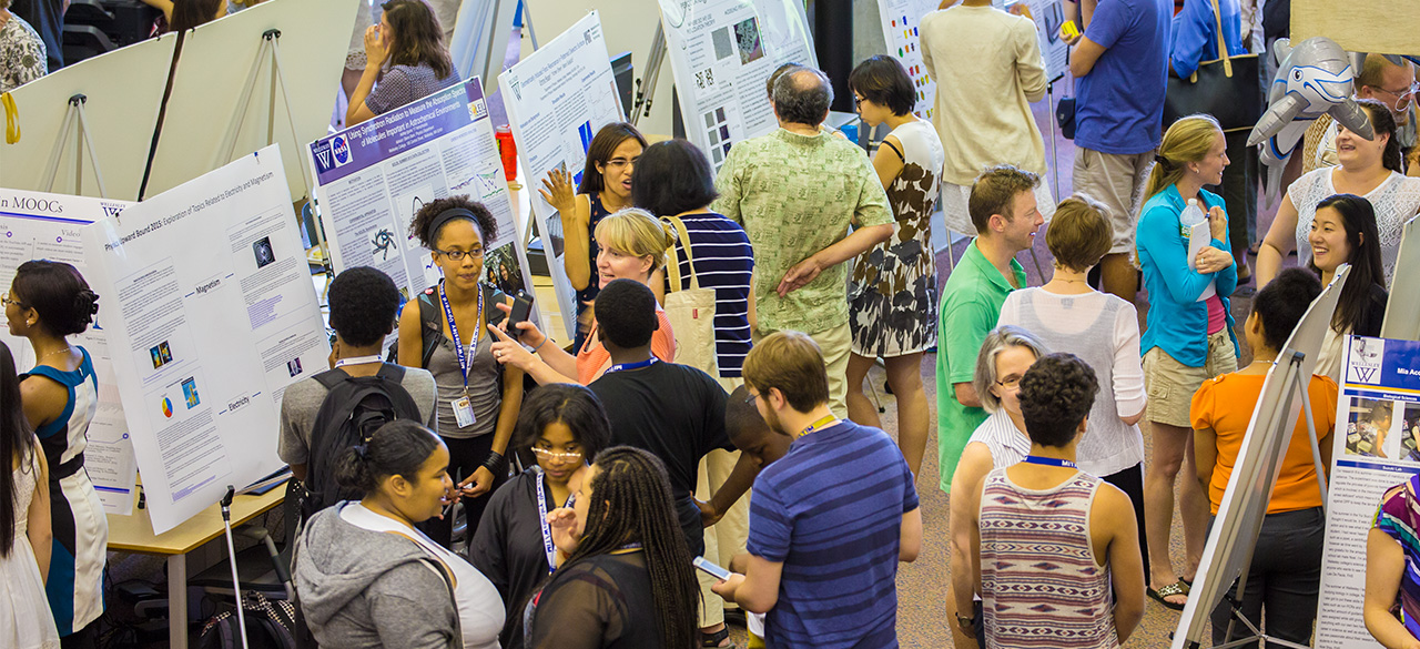 Students, faculty, and visitors gather around posters presenting student summer research.