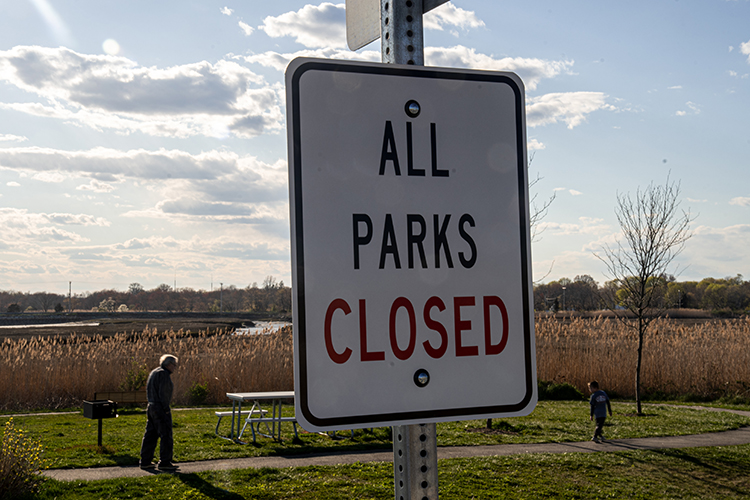 photograph of backlit sign reading ALL PARKS CLOSED, clouds, grass, and reeds in the background