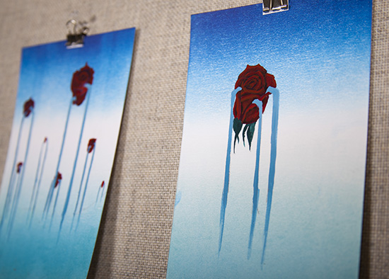 close-up angled photo of 2 small paintings with blue/white gradient backgrounds and solitary dark red roses weeping blue liquid on top