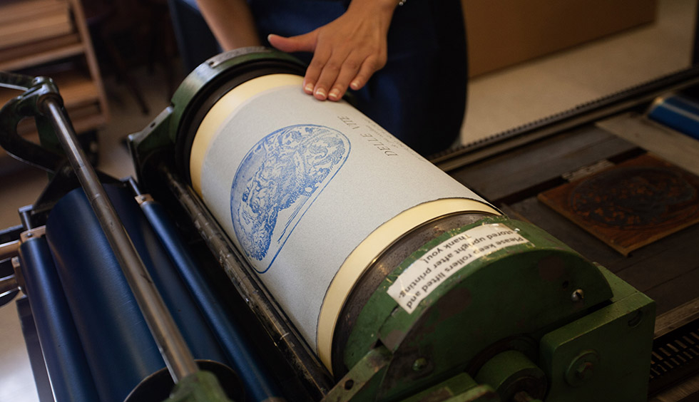 hand holding blue paper with a print of Michelangelo on it to the roller of a press