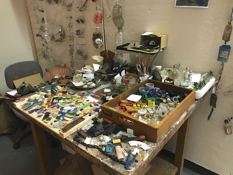 photo of a table in a studio; the entire table surface is covered with bits of trash and boxes full of organized pieces of plastic etc.