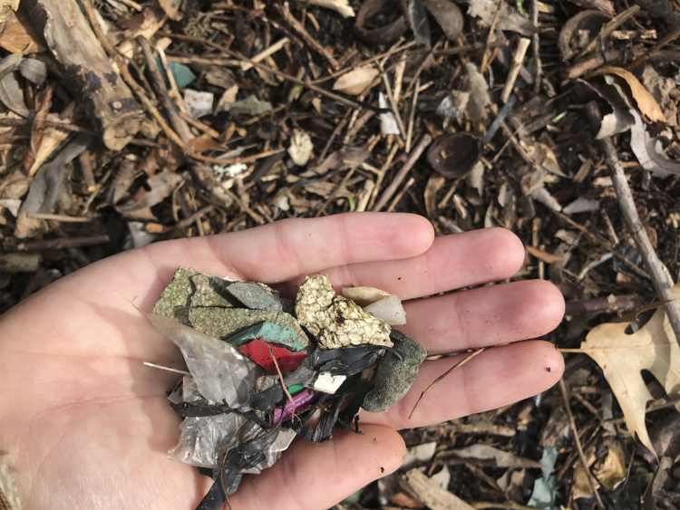 photo of a hand over leaf litter, holding small bits of trash included colorful plastic and dirty styrofoam