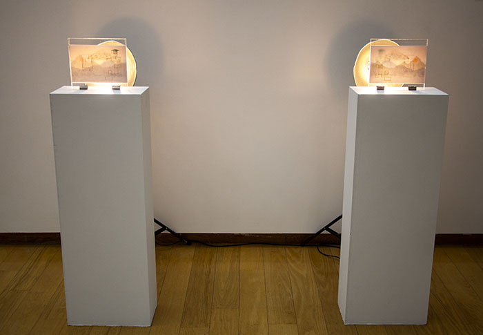 two small layered paper drawings upright on two white pedestals with lights behind