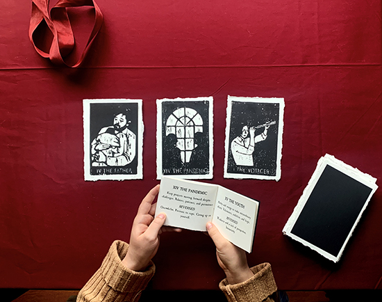 hands holding small book, reads THE PANDEMIC on left page, THE YOUTH on right; three black and white tarot cards on red behind