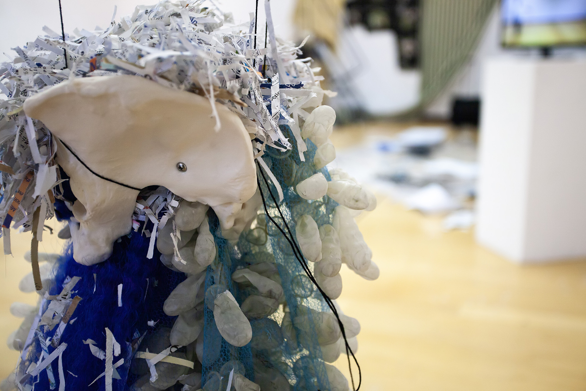 close up on a white pelvis form with shredded paper stuffed in the top, blue tulle, fake hair, and finger cots cascading out of it from below; a messy gallery is visible blurrily behind the form