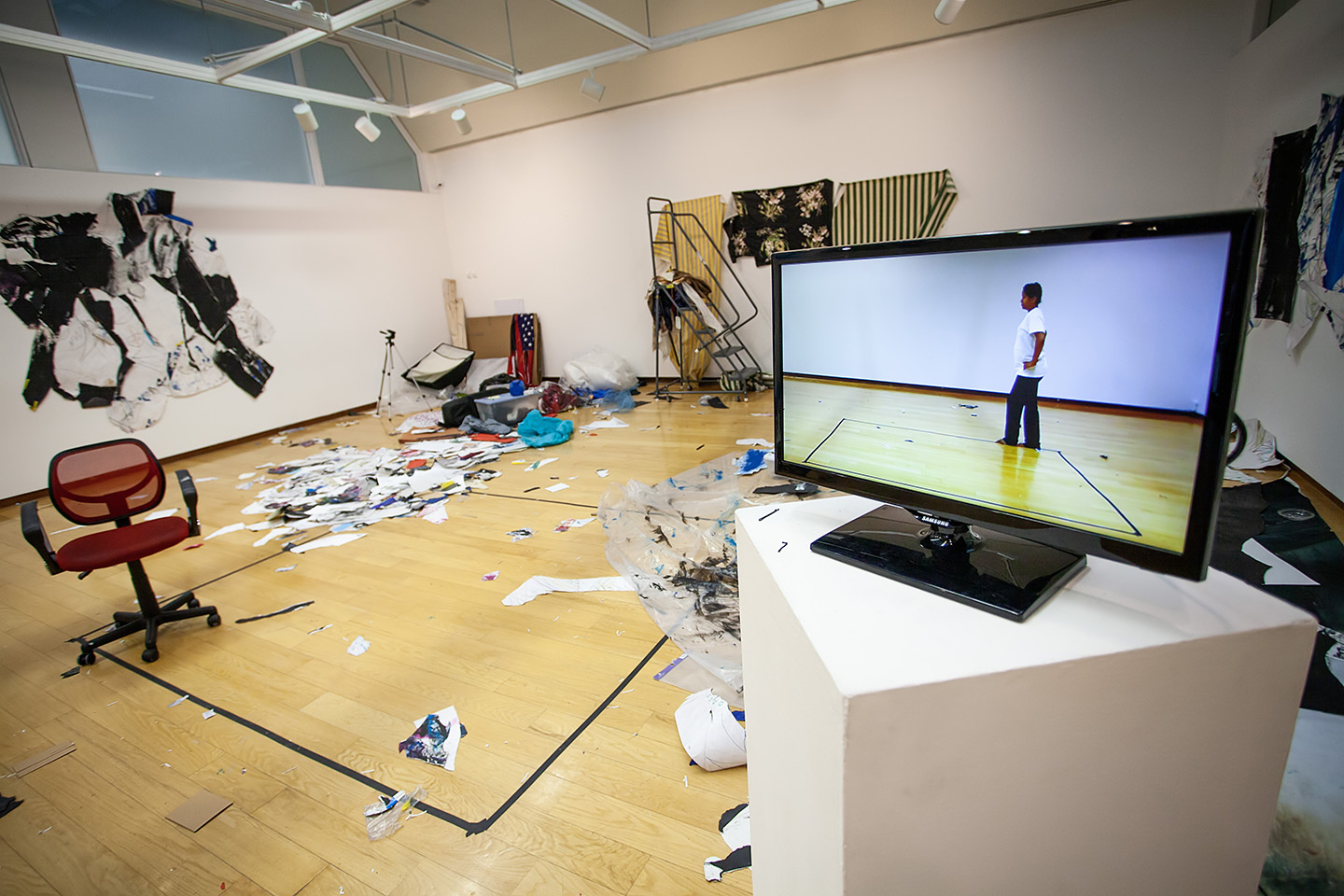 white-walled, wood floor gallery space strewn with piles of paper and other material; on the far wall is a large paper collage, on the back wall three patterned fabrics are hanging; in the foreground, a video screen on a pedestal shows a person walking along a square marked on the floor