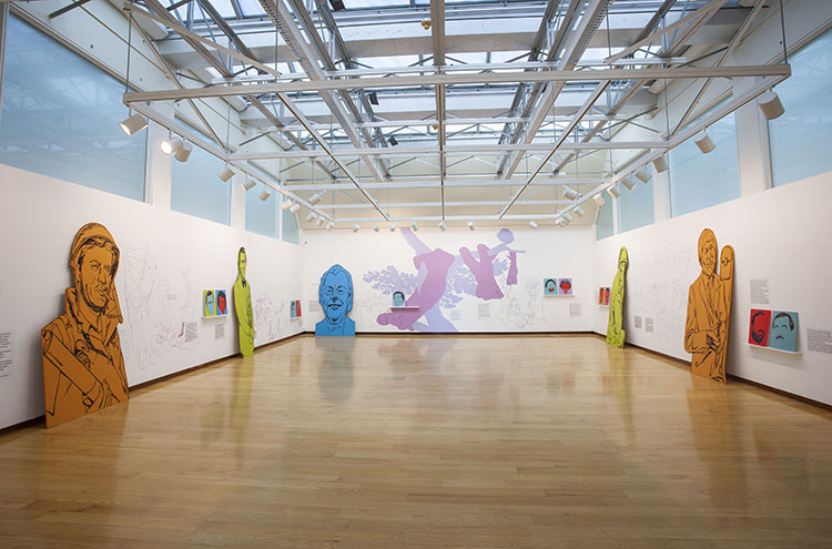 view of gallery with a mix of small paintings, large brightly colored figurative cut-outs, and pink/purple painting directly on the walls