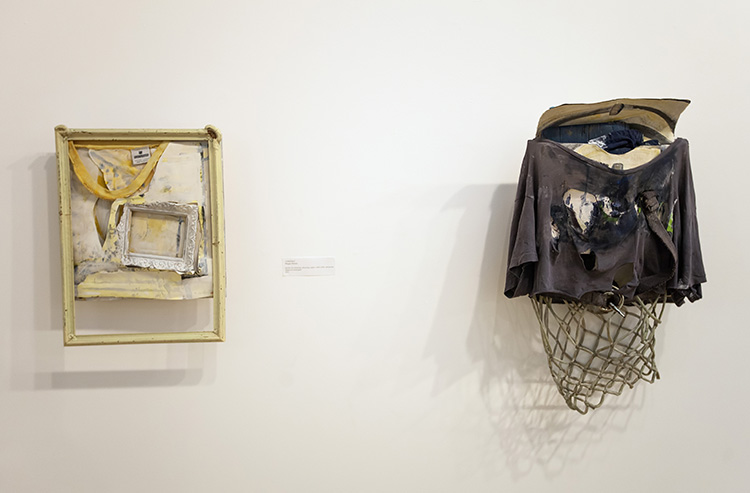 two assemblage works hanging on a wall; at left, yellow-framed piece with fabric scraps and smaller frame inside; at right, paint-covered shirt remnant draped over a boxy form with basketball net hanging below