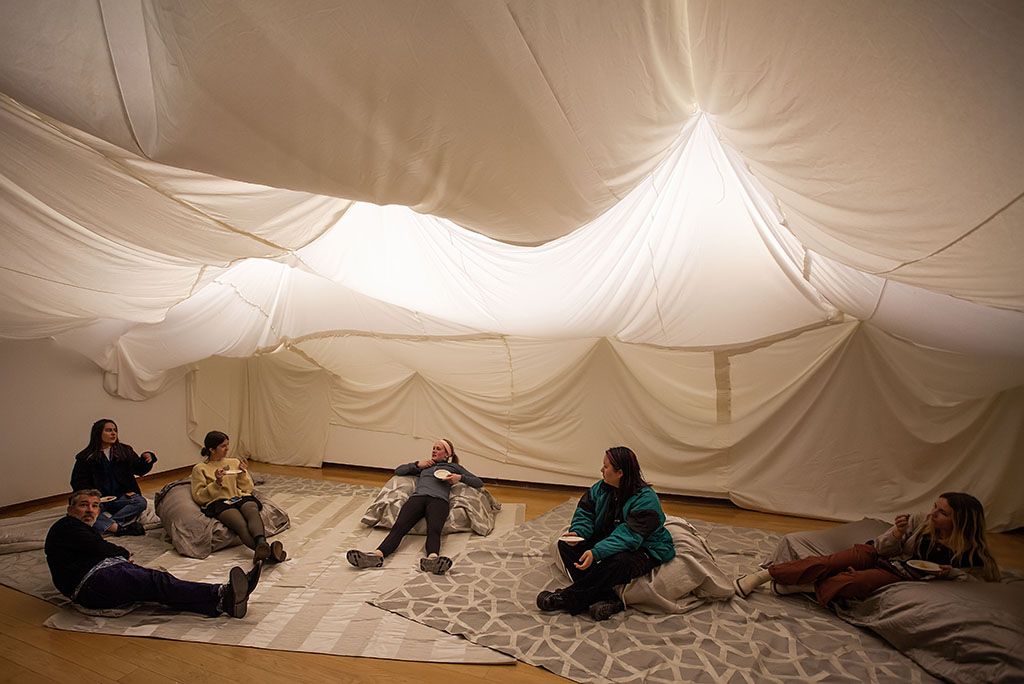 people lounging on scattered gray and white cushions and fabrics, under a low ceiling of variously and dramatically draped white fabric, lit from behind