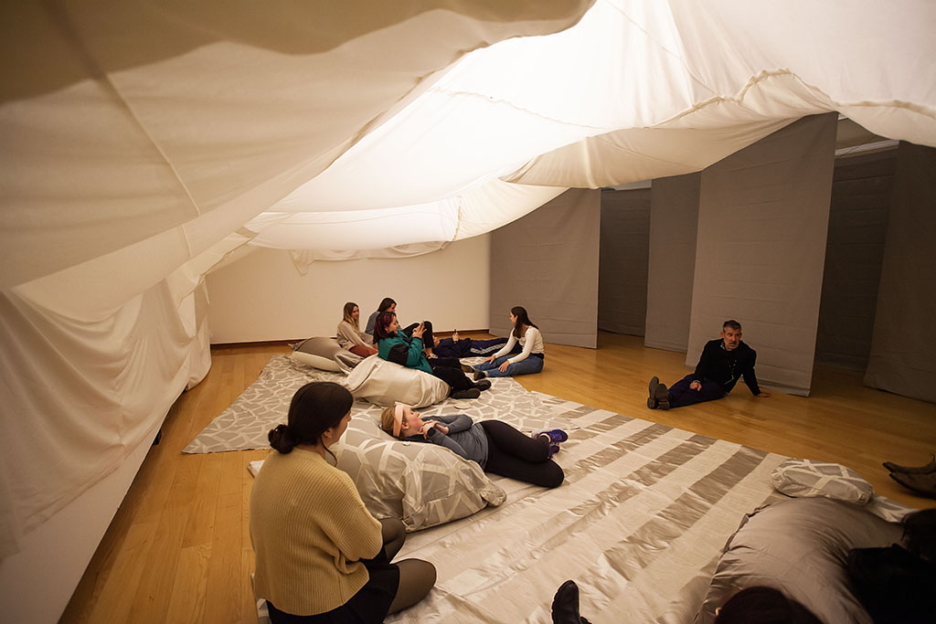 dramatically draped white fabric ceiling, lit from behind, above a group of people sitting on a floor strewn with gray and white fabric and cushions; behind them is a series of gray hanging fabric panels