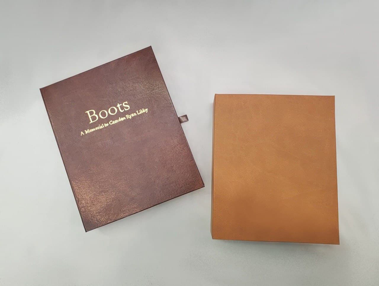 2 artist books lying flat on light gray surface; left book dark red with gold title reading 'Boots: A Memorial to Camden Ryan Libby', right book sienna brown