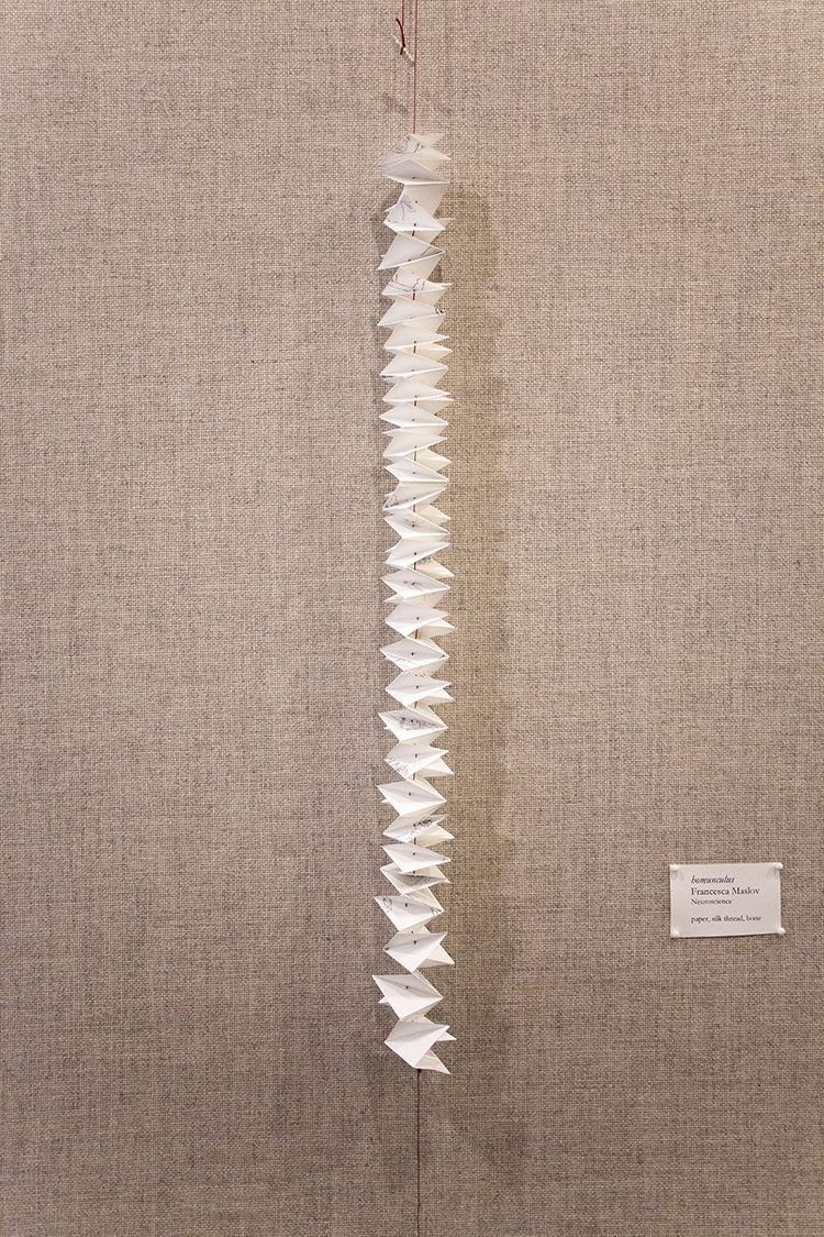 thin column of intensely accordioned paper hanging on a slim red string against a gray fabric wall