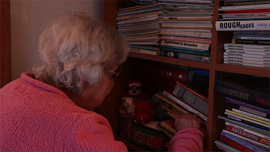 saturated film still of a woman in a pink sweater with white hair at left rummaging in a full bookcase