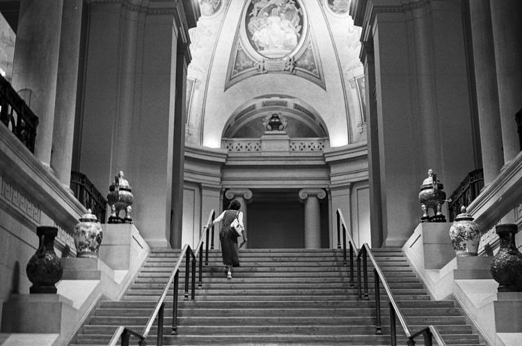 black and white photo of woman walking up museum stairs to large atrium with murals on the walls/ceiling