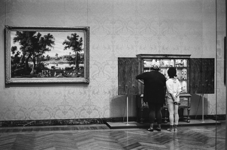 black and white photo of two people looking at cabinet of objects in museum; a large landscape painting is on the wall to their left