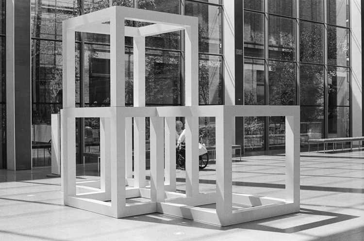 black and white photo of sculpture of open white stacked cubes on exterior museum terrace; patron in wheelchair is partly obscured behind the sculpture