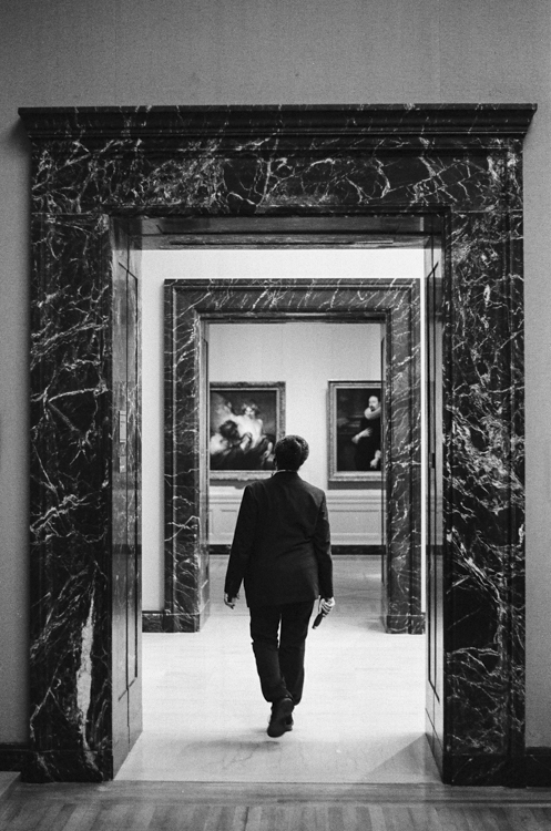 black and white photo of man seen from behind walking through nested marble framed doorways to a room with paintings on the wall