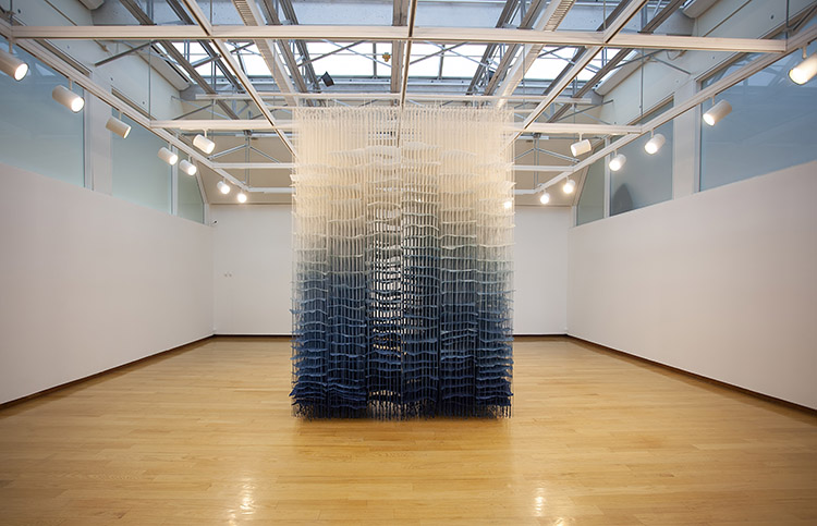 large hanging paper installation, grading from white at the top to blue near the base, hanging in the center of a white-walled, wood floor gallery