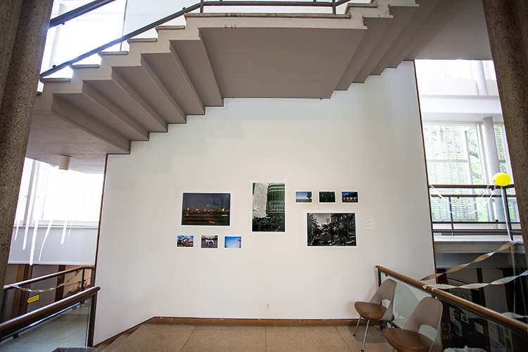 variously sized photographs on a white wall with the underside of stairs above, tiled floor and modernist chairs below