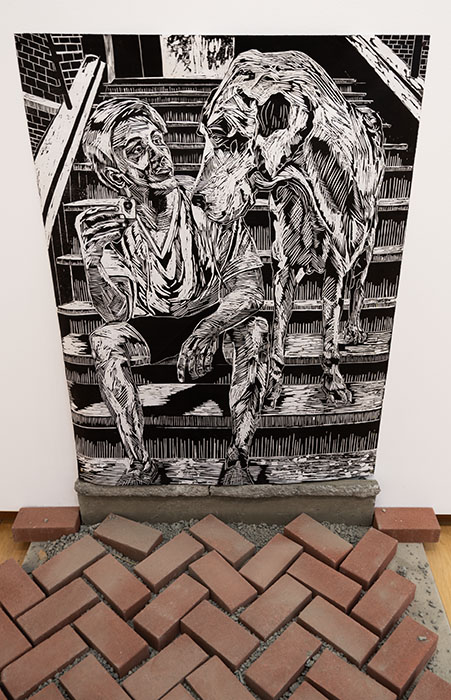 black and white woodcut print of person sitting on steps with a great dane dog, herringbone bricks on the ground in front of the print