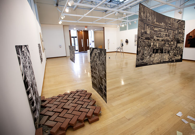 at left, print hung low on wall with bricks arranged in herringbone pattern on floor in front of it, wood panel hanging in front of it; at right, large wood cut panel hanging in the air