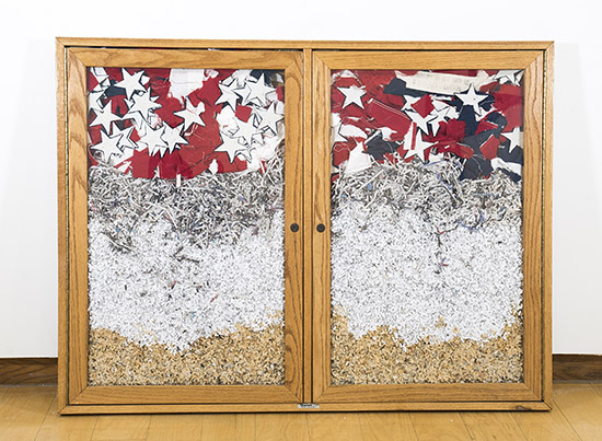 a shallow wall cabinet with two glass doors resting on the floor, stuffed with a shredded American flag and shredded documents