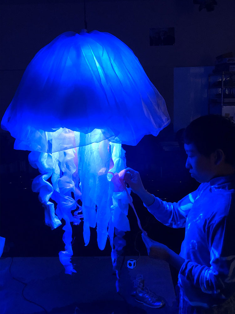 fabric jellyfish lit from within by very intense blue light, with a figure touching its tentacles