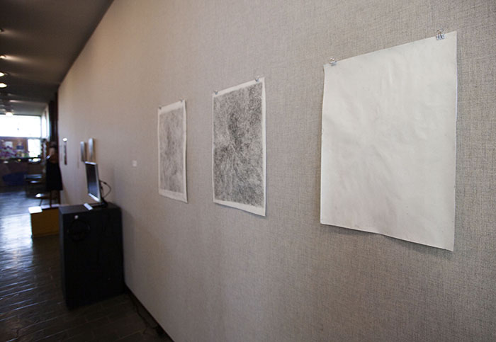 looking from the right at 3 drawings installed on a gray fabric wall, with other artwork beyond them