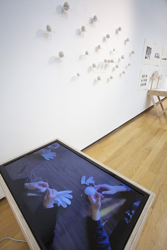 a video of people unraveling gloves displayed flat like a table; behind it is an installation of yarn balls on clear shelves on the wall