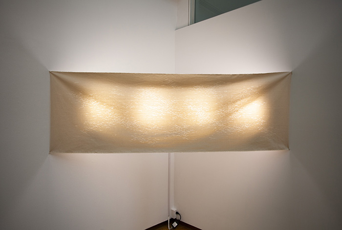 a piece of raw canvas stretched across the corner of a room with white walls. The canvas is pierced with many small lines, through which four lights are shining.
