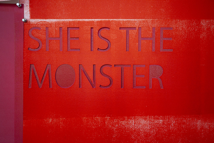 close up of a bright red print with cut-out text reading 'SHE IS THE MONSTER'