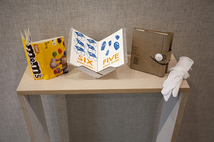Installation of three small handmade books on a small table. The leftmost has a cover made of a yellow M&Ms wrapper. The center is open on a stand showing pages that read 'SIX plastic crates' and 'FIVE plastic bottles'. The rightmost has a cover made from a cardboard Amazon box