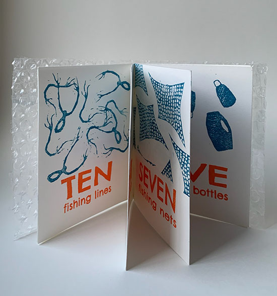 a hand printed book standing up on a table with several pages visible. It has blue ink images of detritus and orange text describing the trash that mimics a child's counting book. The covers are made of bubble wrap.